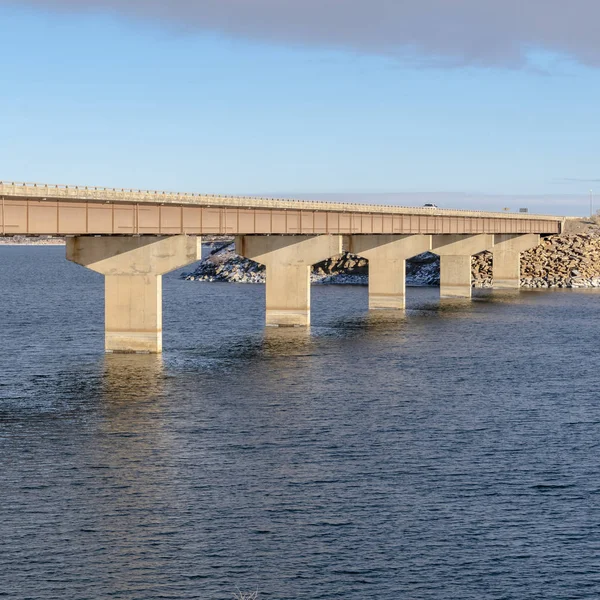 Square Bridge across blue lake with views of snow covered hills and cloudy blue sky — 스톡 사진