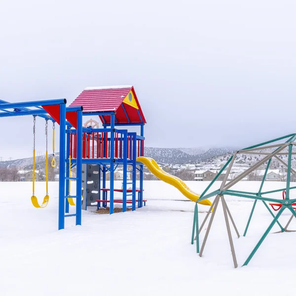 Cornice quadrata Parco innevato con parco giochi contro le case colline e cielo nuvoloso in inverno — Foto Stock