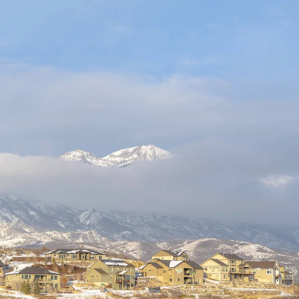 Photo Carré Montagne enneigée partiellement couverte de nuages épais avec des maisons au premier plan — Photo