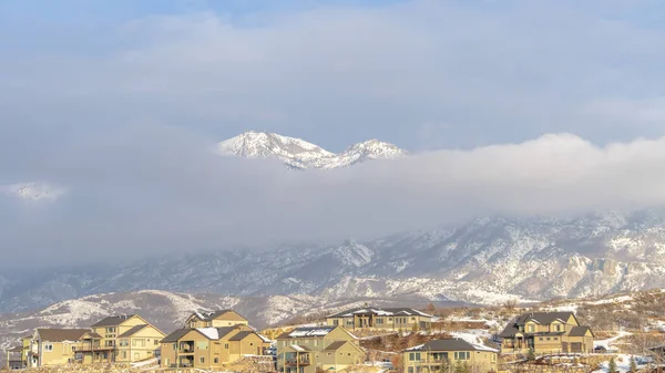 Foto Panorama Montaña nevada parcialmente cubierta de densas nubes con casas en primer plano — Foto de Stock