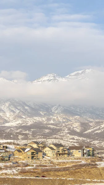 Foto Marco vertical Casas con vista a la montaña nevada parcialmente cubierta de nubes en invierno — Foto de Stock