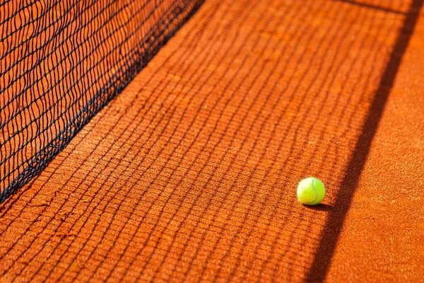 Yellow ball on a clay tennis court next to the net.