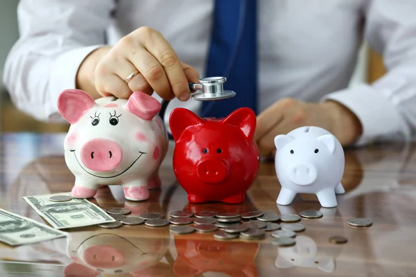 Hand of businessman with stethoscope examining three piggybanks — Stock Photo, Image