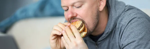 Homem senta-se no sofá e comer hambúrguer contra apartamento — Fotografia de Stock