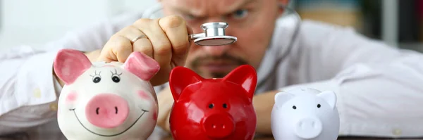 Hand eines Geschäftsmannes mit Stethoskop untersucht drei Sparschweine — Stockfoto