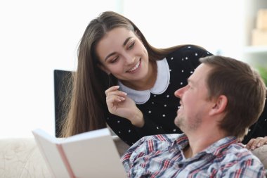 Adam elinde kitapla koltukta oturuyor, kız gülümsüyor.