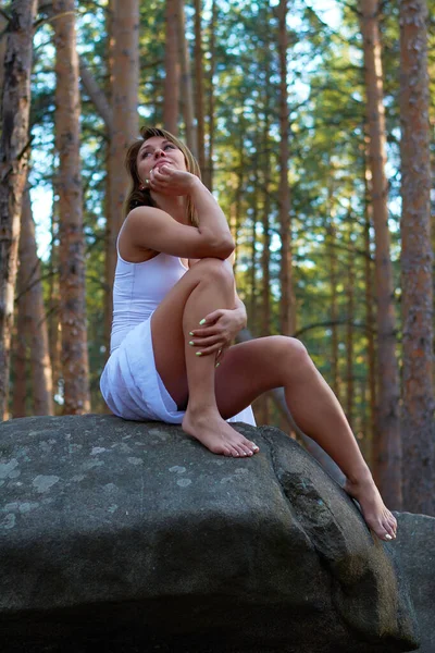 日落时 一个身穿白衣的女孩坐在森林的岩石上 — 图库照片