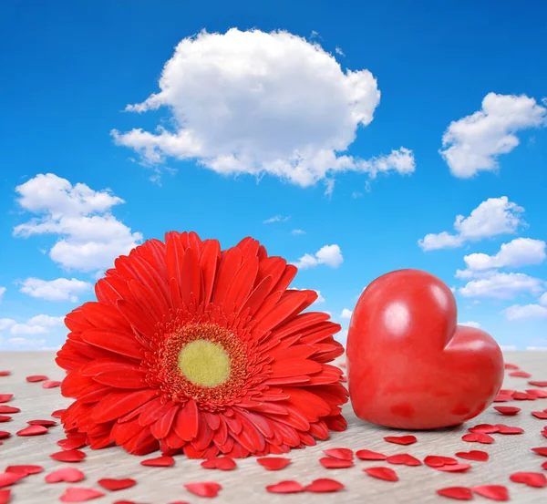 Corazón rojo con flor de gerberas sobre tabla de madera, en el fondo cielo azul con nubes . — Foto de Stock