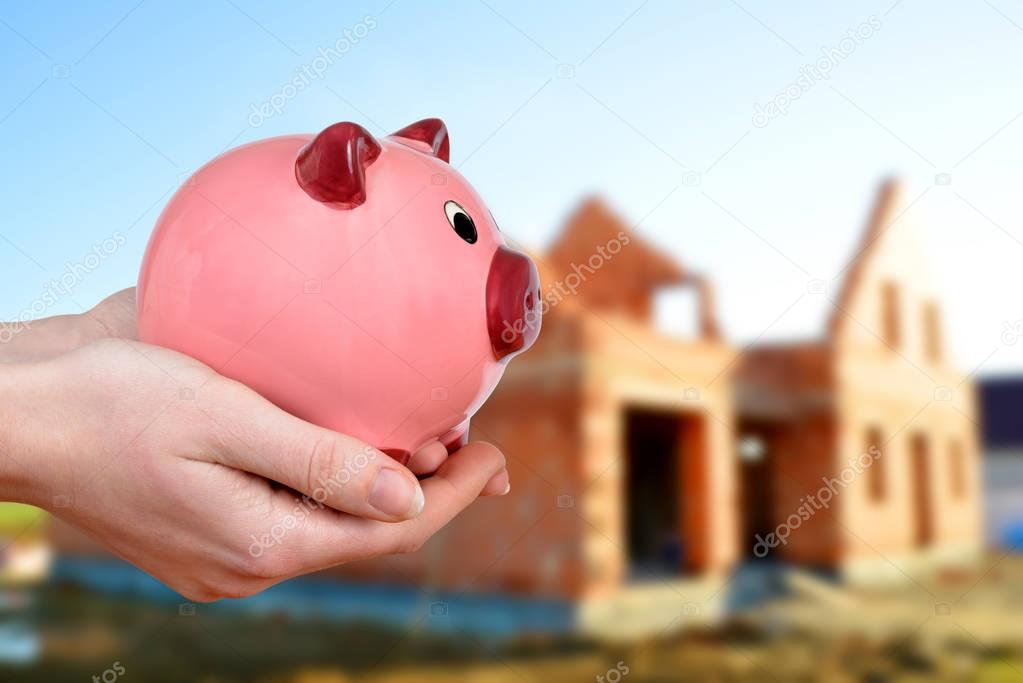 Hands holding pink piggy bank at the background house construction. 