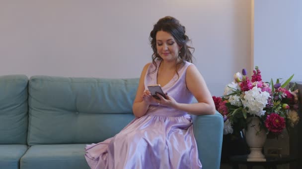 Das Mädchen im Kleid auf der Couch mit dem Handy — Stockvideo