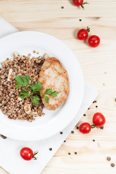 Buckwheat buridge with a cutlet on a white plate on a white cutting board on a wood table with cherry tomatoes and greens — Stok Foto