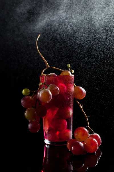 Um copo com bagas de uva, vinho e um monte de uvas em um fundo preto com tambores de gotas — Fotografia de Stock