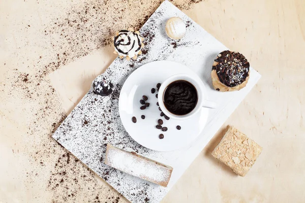 Xícara de café em um pires branco com grãos de café com tortas e bolos em uma placa de corte branca, em um fundo de madeira — Fotografia de Stock