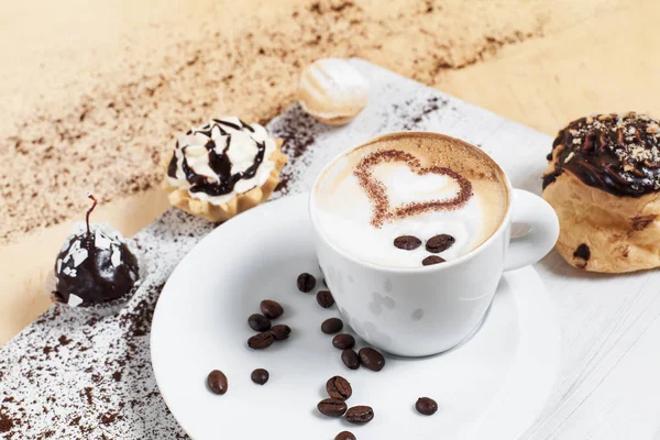 Xícara de café em um pires branco com grãos de café com tortas e bolos em uma placa de corte branca, em um fundo de madeira — Fotografia de Stock