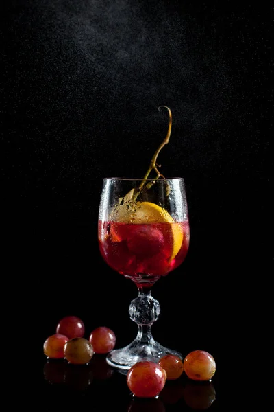 Un bicchiere con acini d'uva, limone, vino e un grappolo d'uva su fondo nero con un filo di gocce — Foto Stock