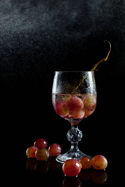 Um copo com bagas de uva, vinho e um monte de uvas em um fundo preto com tambores de gotas — Fotografia de Stock