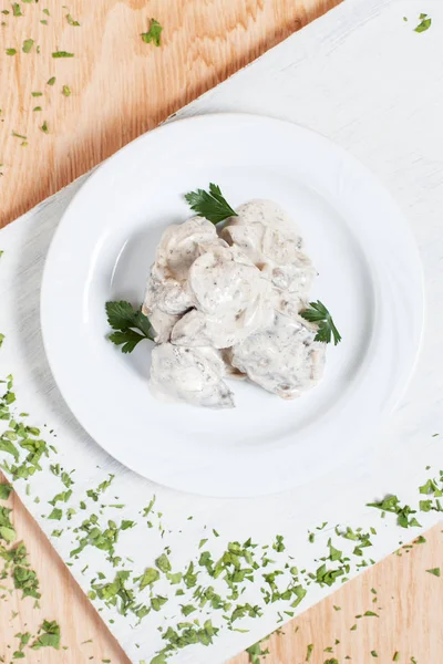 a dish of meat under white sauce on a white plate on a white cutting board with greens on a wooden background