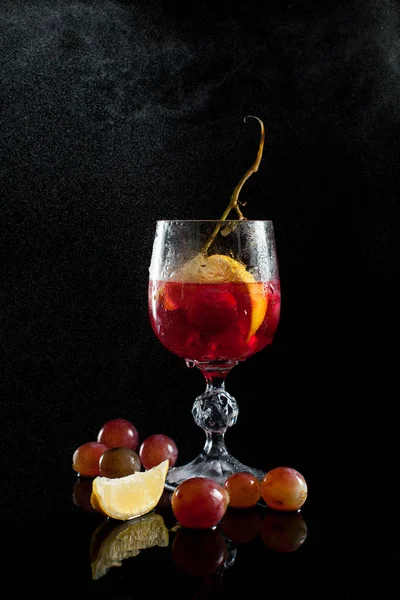 Un bicchiere con acini d'uva, limone, vino e un grappolo d'uva su fondo nero con un filo di gocce — Foto Stock