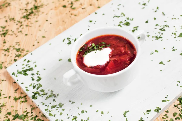 Свекловичный суп с овощами, травами и мясом в белой миске, на разделочной доске на деревянном столе — стоковое фото