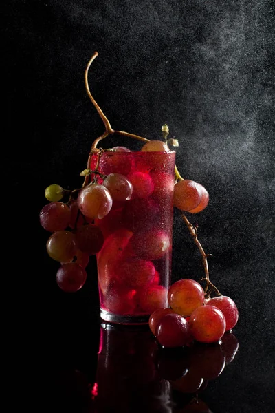 Un bicchiere con acini d'uva, vino e un grappolo d'uva su fondo nero con fusti di gocce — Foto Stock