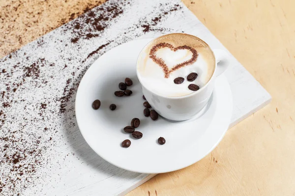 Taza de café en un platillo blanco con granos de café con pasteles y pasteles en una tabla de cortar blanca, sobre un fondo de madera — Foto de Stock