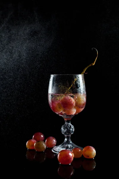 Um copo com bagas de uva, vinho e um monte de uvas em um fundo preto com tambores de gotas — Fotografia de Stock
