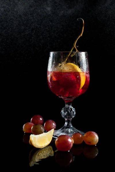 Un bicchiere con acini d'uva, limone, vino e un grappolo d'uva su fondo nero con un filo di gocce — Foto Stock