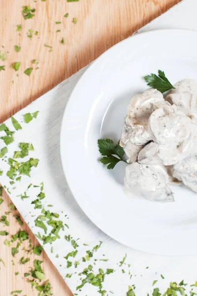 a dish of meat under white sauce on a white plate on a white cutting board with greens on a wooden background