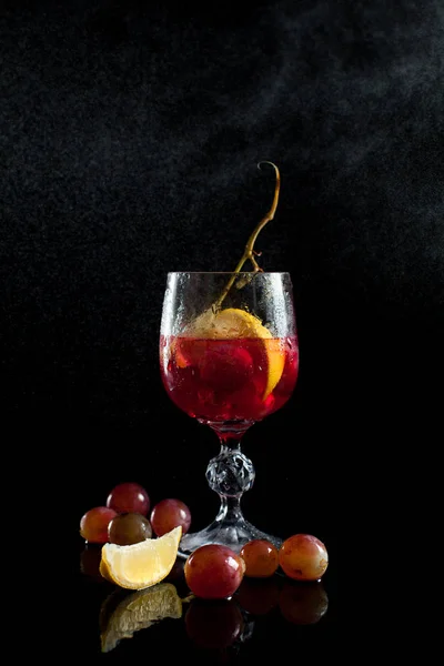 Un verre avec des baies de raisin, citron, vin et un bouquet de raisins sur un fond noir avec un filet de gouttes — Photo