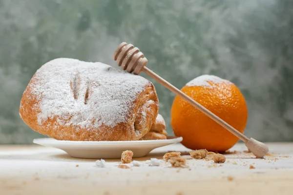bir puf kek meyve doldurma ve çikolata beyaz tabakta ile pudra şekeri serpilir ve çikolata bir ahşap tatlım sopa ve turuncu ile mavi bir arka plan üzerinde ahşap bir masaya döktü