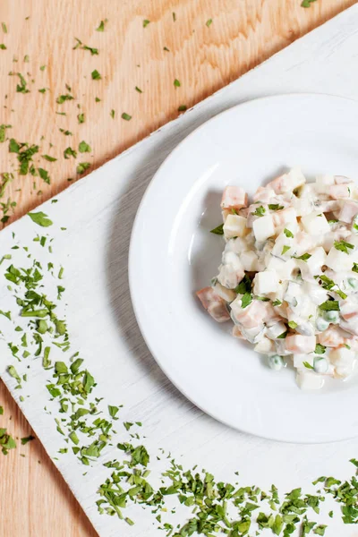 Insalata Olivier con verdure su un piatto bianco su un asciugamano su un tagliere bianco su uno sfondo di legno — Foto Stock