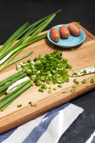 green onion in the process of slicing with a knife with a plate of sausages on a wooden cutting board and a kitchen towel on a dark background
