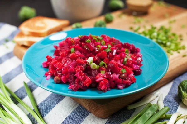 Салат со свеклой, тост с сыром, зеленый лук и миска с брюссельской капустой на деревянной доске, полосатое кухонное полотенце на темном деревянном фоне — стоковое фото