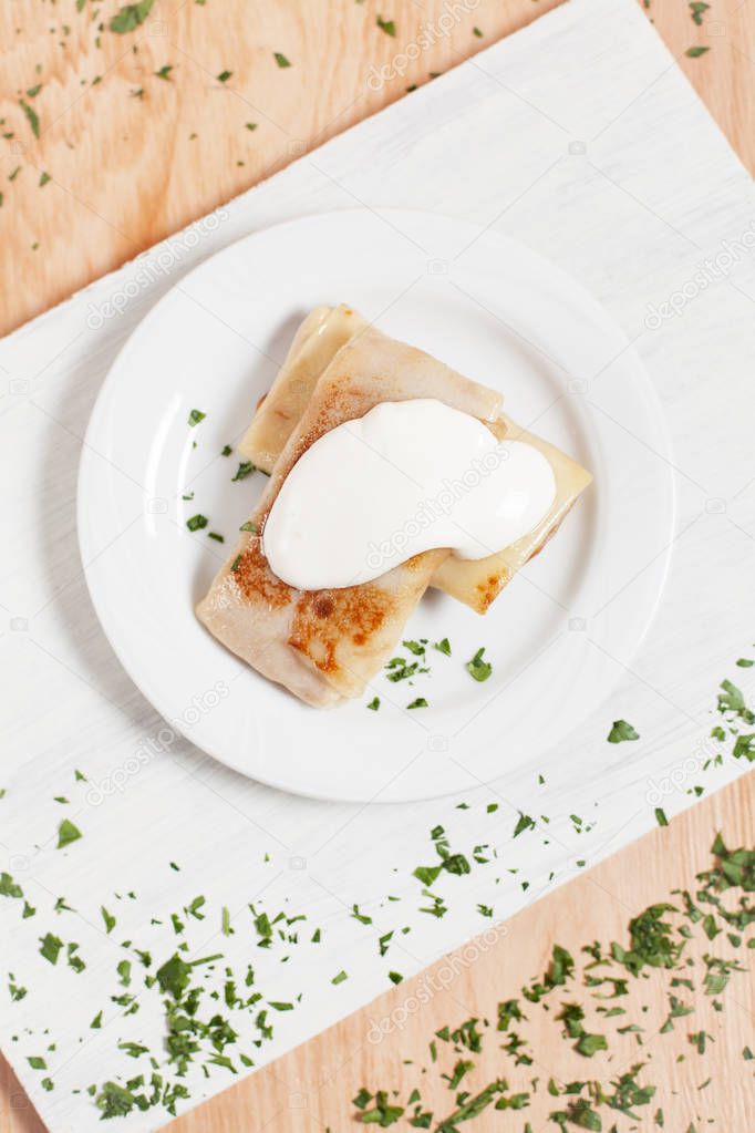 pancakes with filling and sour cream on a white plate on a white cutting board on a light wooden background sprinkled with herbs