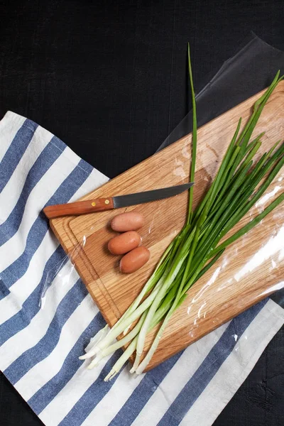 green onion in the process of slicing with a knife with sausages on a wooden cutting board and a kitchen towel on a dark background