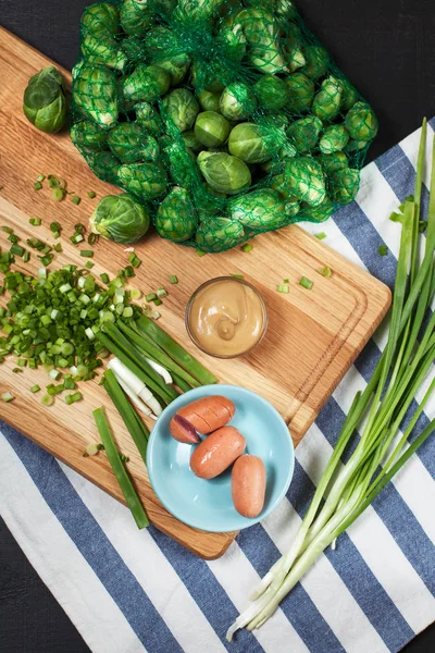 green onion in the process of slicing with sausages on a blue saucer and Brussels sprouts on a wooden cutting board and a kitchen towel on a dark background