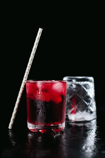 Cocktail rosso con ghiaccio e bicchiere vuoto, con una sfumatura su fondo nero bagnato e un tubo — Foto Stock