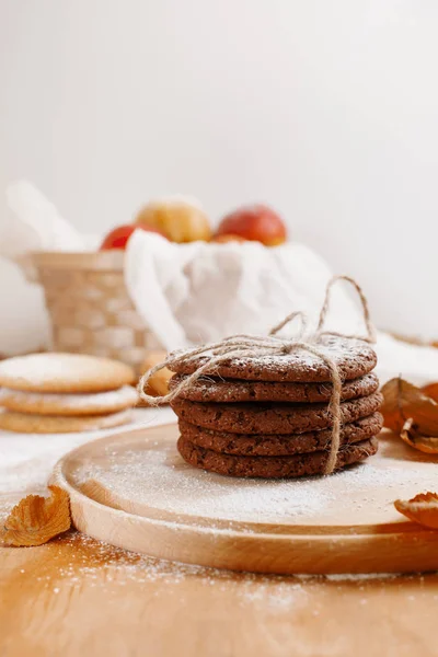 round cookies with sugar powder on a round wooden board, a basket of apples, a white towel on a light background surrounded by autumn leaves