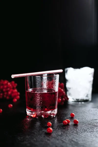 Bicchiere con cocktail rosso e paglia, bicchiere con ghiaccio e bacche rosse su fondo nero bagnato — Foto Stock