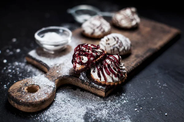 Small chocolate cakes with different stuffings, sprinkled with berry topping, sprinkled with powdered sugar on a wooden board on a dark background — Stock Photo, Image