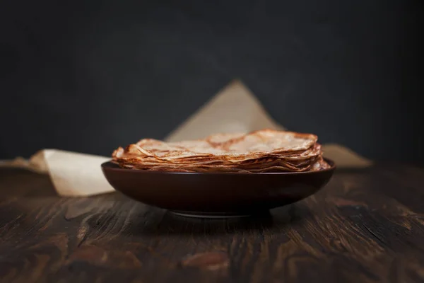 Koyu gri arka plan üzerinde koyu ahşap bir masa üzerinde kahverengi bir tabak içinde yığılmış krep — Stok fotoğraf