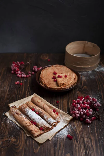 Sweet fresh pancakes with a pile and pancakes on baking paper, rolled with tubules, viburnum berries on a dark wooden table on a dark gray background — Stock Photo, Image
