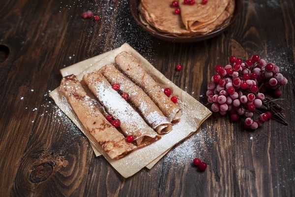 Sweet fresh pancakes with a pile and pancakes on baking paper, rolled with tubules, viburnum berries on a dark wooden table on a dark gray background — Stock Photo, Image