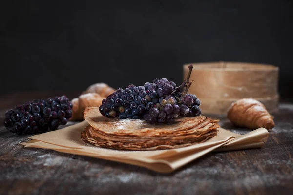 Panqueques dulces recién horneados apilados sobre papel de hornear marrón, croissants y uvas sobre una mesa de madera marrón oscuro sobre un fondo gris oscuro — Foto de Stock
