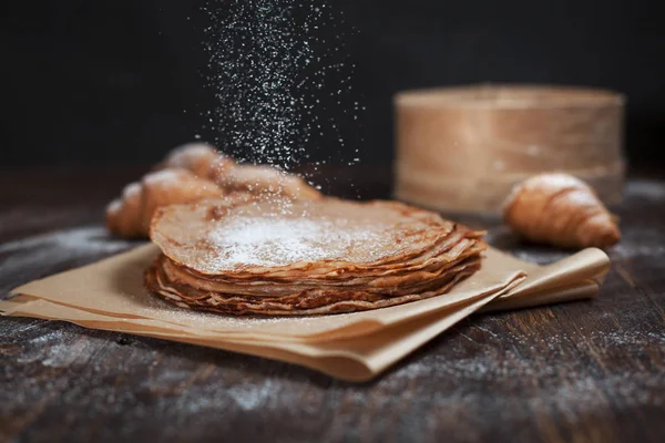 Freshly baked sweet pancakes stacked on brown baking paper and croissants on a dark brown wooden table on a dark gray background — Stock Photo, Image
