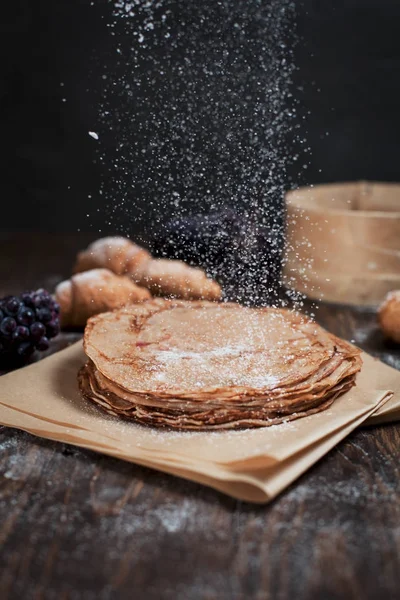 Freshly baked sweet pancakes stacked on brown baking paper, croissants and grapes on a dark brown wooden table on a dark gray background — Stock Photo, Image