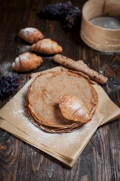 Panqueques dulces recién horneados apilados sobre papel de hornear marrón, croissants y uvas sobre una mesa de madera marrón oscuro sobre un fondo gris oscuro — Foto de Stock