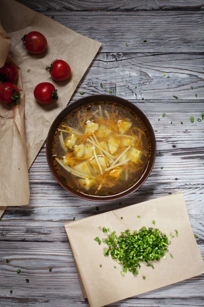 Zuppa vegetariana di verdure fresche, verdure e pasta in un piatto marrone, cipolla verde tritata su carta da forno marrone e pomodorini su un tavolo di legno leggero — Foto Stock