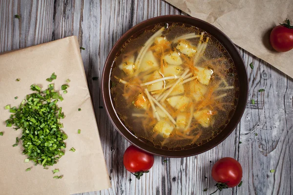 Zuppa vegetariana di verdure fresche, verdure e pasta in un piatto marrone, cipolla verde tritata su carta da forno marrone e pomodorini su un tavolo di legno leggero — Foto Stock