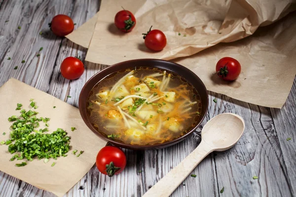 Vegetarisk Soppa av färska gröna, grönsaker och pasta i en brun tallrik, hackad grön lök på brunt bakplåtspapper och körsbärstomater på ett ljust träbord — Stockfoto